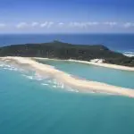 Stunning aerial view of Australia’s longest surfable wave at a sandy beach near a lush, forested island—top surfing destination.