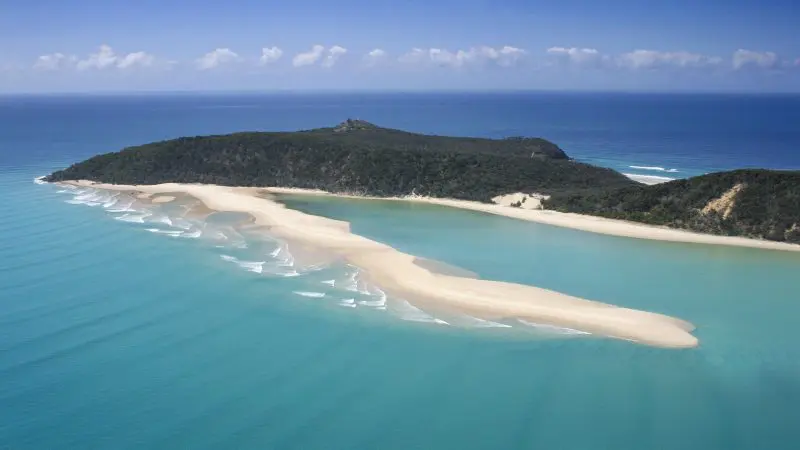 Stunning aerial view of Australia’s longest surfable wave at a sandy beach near a lush, forested island—top surfing destination.