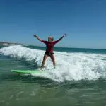 Surfer in a red shirt rides a gentle wave at Learn To Surf Australia’s Longest Wave, arms wide open under clear blue skies.