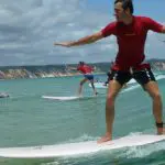Man in red shirt rides a small wave at Learn To Surf Australia's Longest Wave, scenic cliffs and fellow surfers visible behind him.