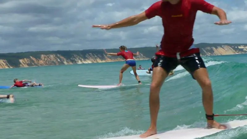 Man in red shirt rides a small wave at Learn To Surf Australia's Longest Wave, scenic cliffs and fellow surfers visible behind him.