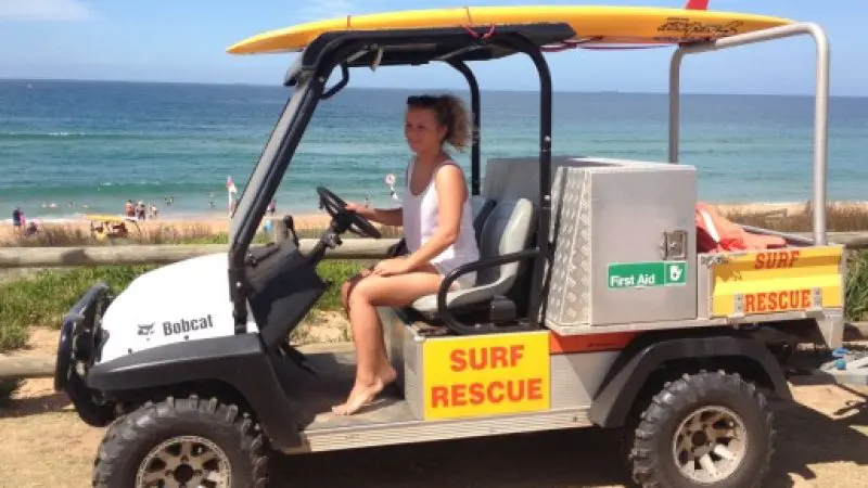 Woman in Celebtime Tours surf rescue vehicle with surfboard on top, parked by sunny beach—ideal for lifeguard services and tours.