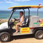 Woman enjoying a sunny Celebtime 1 Day Home Away Location Tour, seated in a surf rescue vehicle with yellow surfboard by the beach.