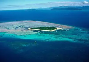 Stunning aerial view of a lush green island encircled by vibrant turquoise waters, colourful coral reefs, and expansive deep blue ocean.