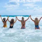 Four women in swimming costumes hold hands, enjoying ocean waves on The Great Escape 21 Day Trip from Brisbane to Cairns, creating lasting memories.