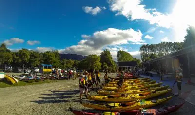 Participants in bright life jackets assemble by vibrant yellow kayaks for the Three Day Classic under clear blue skies amid lush trees.