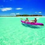 Adventurers kayaking through crystal-clear waters towards a secluded island on a 1 Day K'gari Fraser Island tour beneath sunny blue skies.