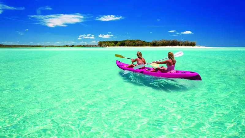 Adventurers kayaking through crystal-clear waters towards a secluded island on a 1 Day K'gari Fraser Island tour beneath sunny blue skies.