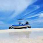 A blue inflatable boat on pristine white sand awaits a Remote 1 Day K'gari Fraser Island tour beneath a vivid, inviting sky.