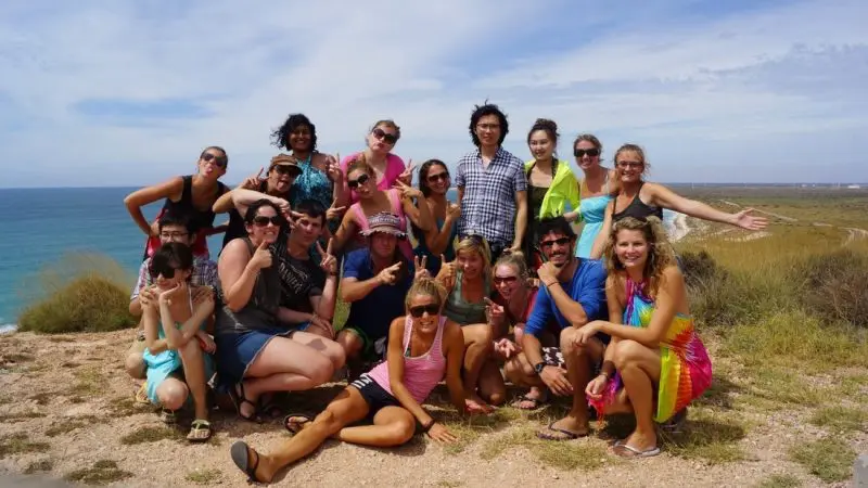 A smiling tour group on a 6 Day Perth to Exmouth adventure stands atop a scenic coastal cliff with sweeping ocean and coastline views.