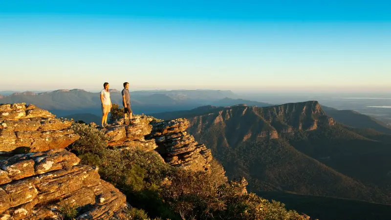 Two travellers stand atop a rugged cliff, admiring sweeping Grampians mountain vistas under a bright blue sky on their day tour.