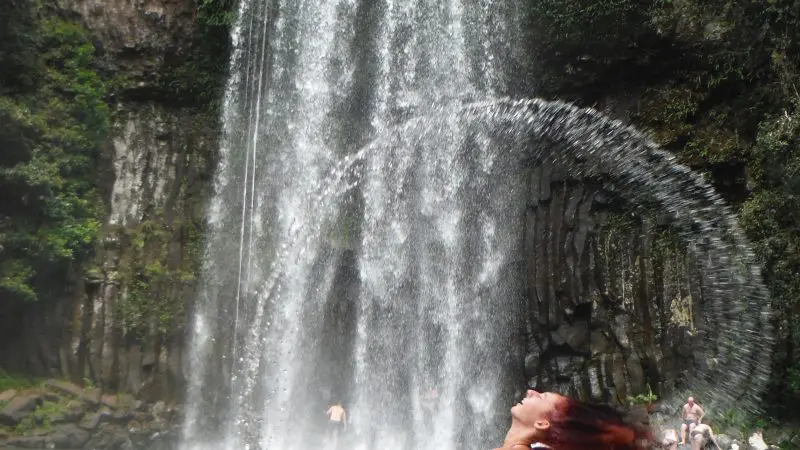 Woman flicks her hair in a sparkling waterfall pool on a Daintree Rainforest tour, water arc glistening, swimmers visible behind.
