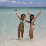 Two women in matching blue bikinis joyfully celebrate in crystal-clear sea water on a Remote 1 Day K'gari Fraser Island tour experience.