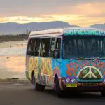 Vibrant, hippy-style Happy Coach Byron Bay bus parked on a scenic beach at sunset with mountains in the background.