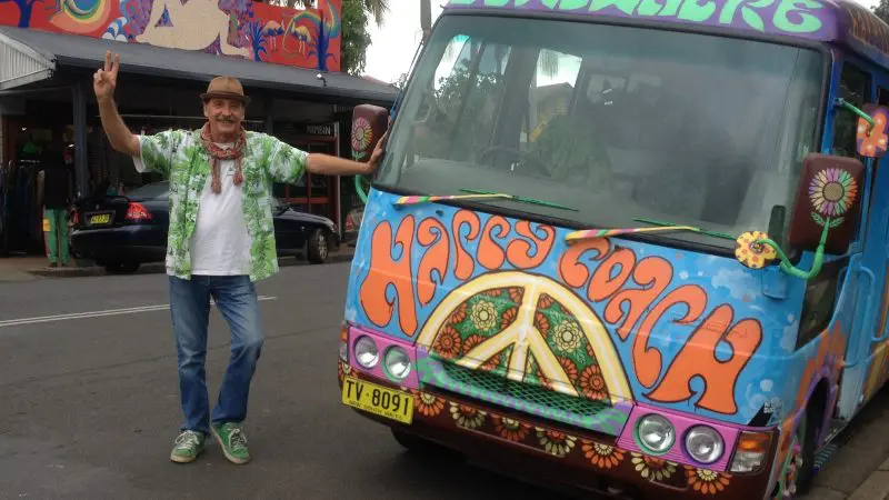 A traveller stands next to a vibrant, psychedelic Happy Coach bus on Byron Bay’s 1 Day Nimbin Tour, amid lush palm trees.