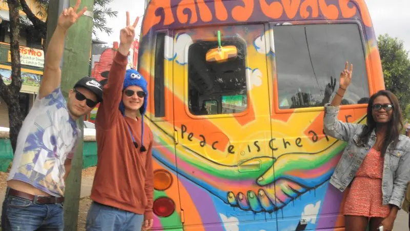 Three people smiling and flashing peace signs beside the colourful Happy Coach van during a 1 Day Nimbin Tour from Byron Bay.