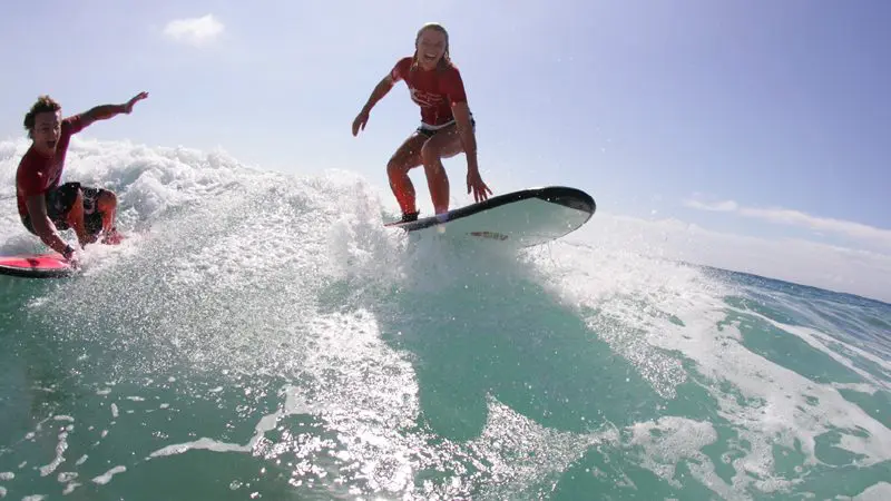 Experience the thrill as two surfers in red catch a wave beneath clear skies on the Byron Bay Learn to Surf Half-Day Tour.