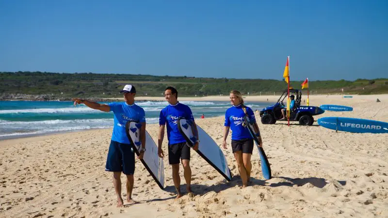 Three surfers in matching blue shirts carry surfboards along Byron Bay beach after a one-and-a-half-hour private Let’s Go Surfing lesson, sunny day.