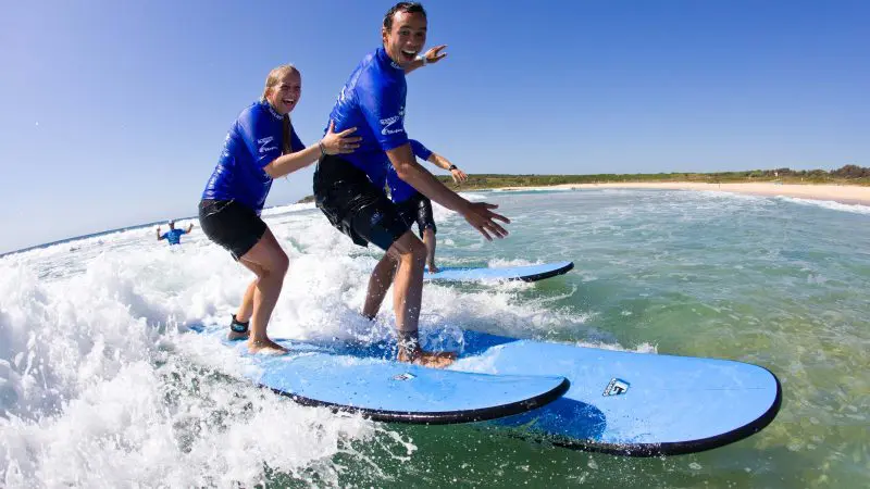 Two surfers in blue shirts catch gentle waves close to shore during a Byron Bay 2 Hour Small Group Surf Lesson for beginners.