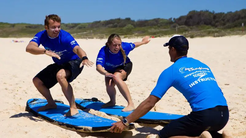 Join a Byron Bay 2 Hour Small Group Surf Lesson, as two people master surf stances on sand, preparing to ride real waves confidently.
