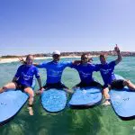 Smiling group in matching blue shirts pose confidently on surfboards under sunny skies during a Byron Bay Learn to Surf Half Day Tour.