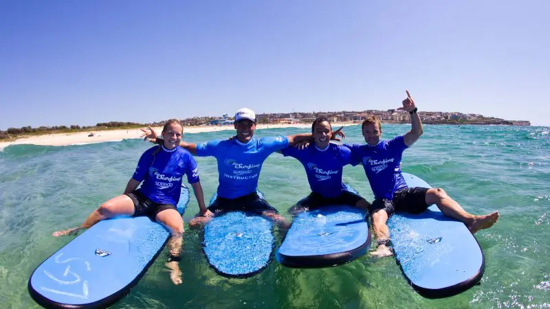 Smiling group in matching blue shirts pose confidently on surfboards under sunny skies during a Byron Bay Learn to Surf Half Day Tour.