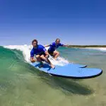 Two surfers in blue shirts ride gentle waves at Byron Bay during a two-hour small group surf lesson under clear, bright blue skies.