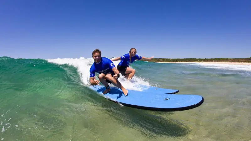 Byron Bay surf lesson: two people in blue shirts riding small waves on blue boards, Learn to Surf Half-Day Tour experience.
