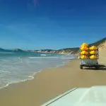 Yellow Dolphin Kayaking gear on a trailer towed down Rainbow Beach, bordered by turquoise ocean waves and dramatic rocky cliffs.
