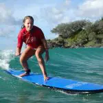 Young woman in red shirt surfing near coastal rocks at Australia’s Longest Wave Beach Drive by Learn To Surf—expert surf lessons.