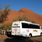 White Adventure Tours coach for 3-Night Uluru Rock trip parked by iconic red rock formations; travellers explore the area nearby.