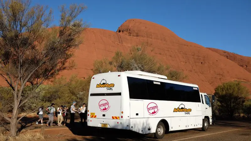 White Adventure Tours coach for 3-Night Uluru Rock trip parked by iconic red rock formations; travellers explore the area nearby.