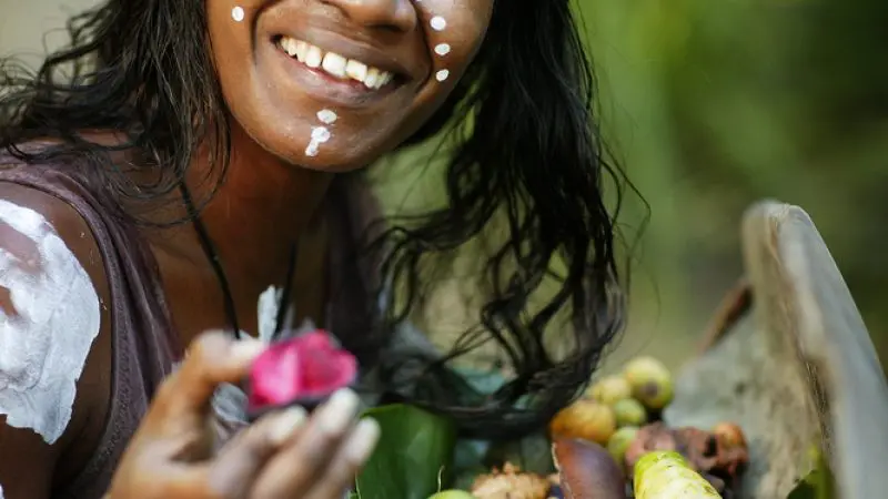 A joyful woman with white face paint savours native fruits and roots atop green leaves, capturing the essence of the Great Australian Wildlife Experience.