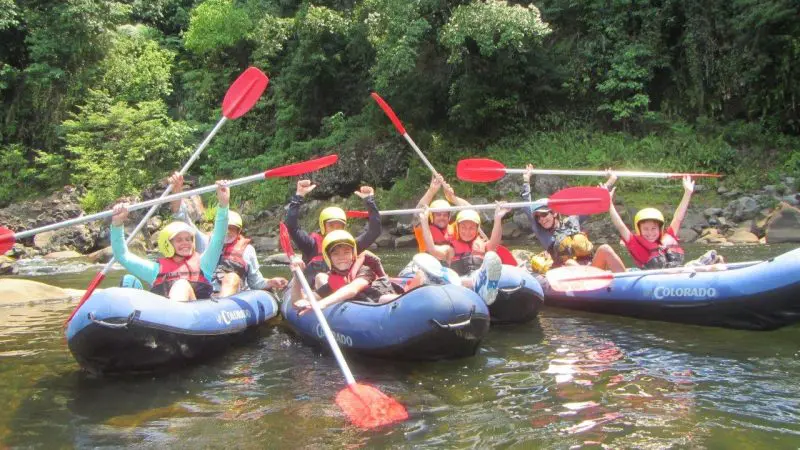 Thrilled group on Sports Rafting Full Day Ex Mission Beach, paddles raised high, smiling faces, surrounded by lush rainforest scenery.