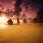Breathtaking Great Ocean Road sunset: golden sandy beach, towering limestone rock stacks, footprints in soft sand, moody clouded sky.