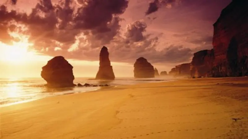 Breathtaking Great Ocean Road sunset: golden sandy beach, towering limestone rock stacks, footprints in soft sand, moody clouded sky.