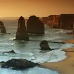 Majestic rock formations emerge from turquoise sea waters beside dramatic cliffs on a Great Ocean Road sunset tour, golden sands below.