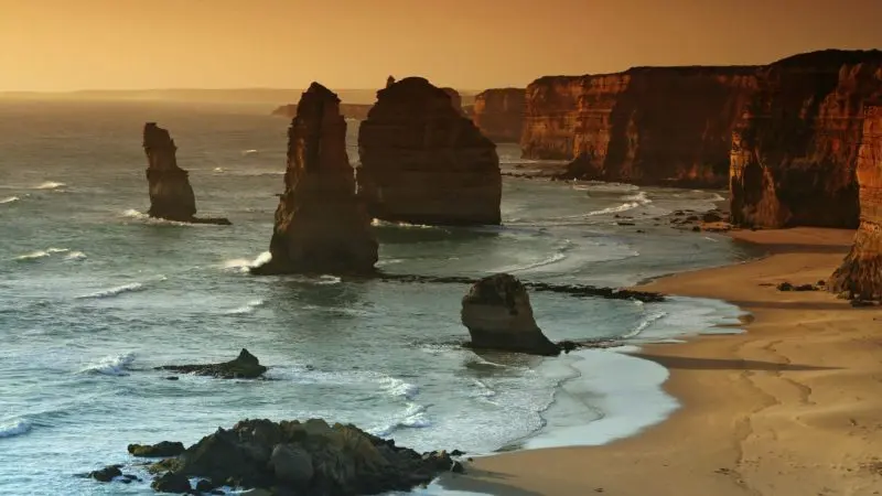 Majestic rock formations emerge from turquoise sea waters beside dramatic cliffs on a Great Ocean Road sunset tour, golden sands below.