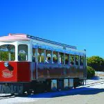Red and white open-air tram on Rottnest Island’s sandy track, passing lush green bushes under blue sky—Oliver Hill Train Tunnel Tour.