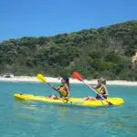 Kayakers paddle on crystal-clear blue water by Rainbow Beach’s lush green hills, enjoying a top-rated Great Beach Drive adventure.