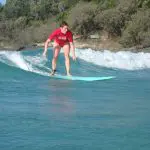 Surfer in vibrant red shirt riding Australia’s Longest Wave by a pristine, sandy, tree-lined shore under a bright blue sky.