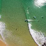 Aerial shot of surfers catching waves on Australia’s Longest Wave, with crystal-clear waters and pristine sandy beach in view.