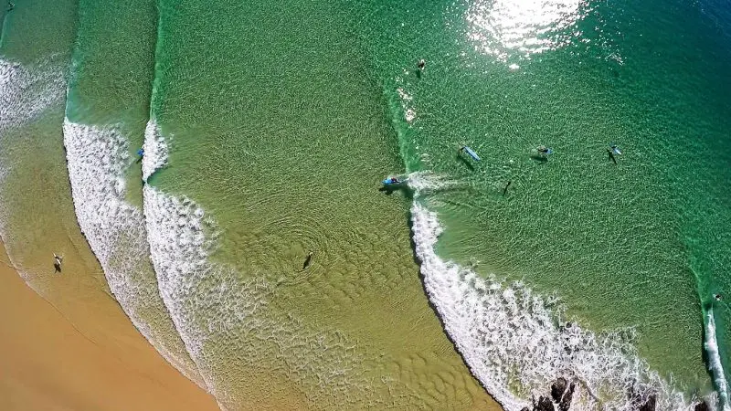 Aerial shot of surfers catching waves on Australia’s Longest Wave, with crystal-clear waters and pristine sandy beach in view.