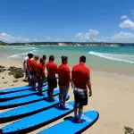 Surf lesson at Australia's Longest Wave Beach Drive: group in red shirts on blue surfboards faces sea, Learn To Surf school.