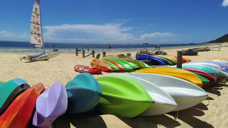 Vibrant kayaks rest on sandy Moreton Island shores, ready for adventure with a sailboat and clear blue sky in the background.