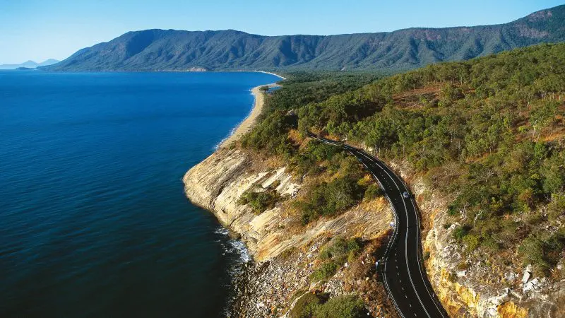 Scenic coastal road curves beside turquoise sea and lush green hills on the Cape Tribulation Daintree Wilderness Tour in Australia.