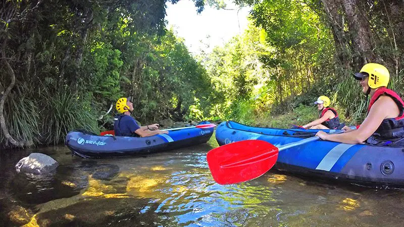 Three adventurers in helmets paddle blue inflatable kayaks on a Half Day Sports Rafting tour along a scenic, tree-lined clear stream.