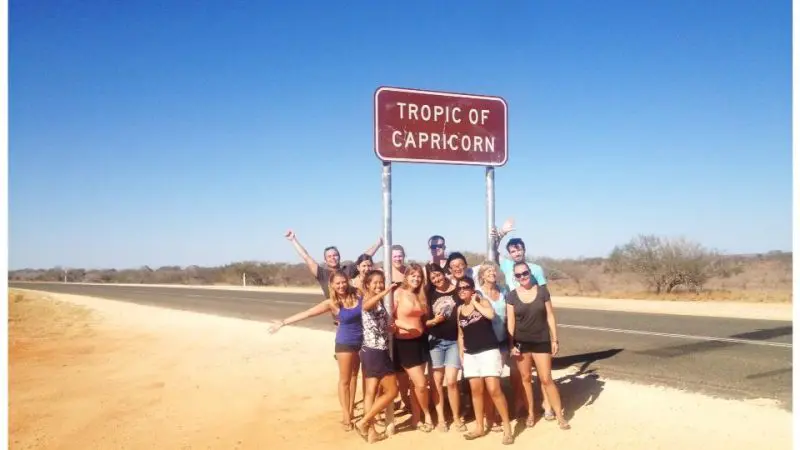 Smiling group on 6 Day Perth to Exmouth One Way Tour posing beneath Tropic of Capricorn sign in Australia’s sunlit outback scenery.