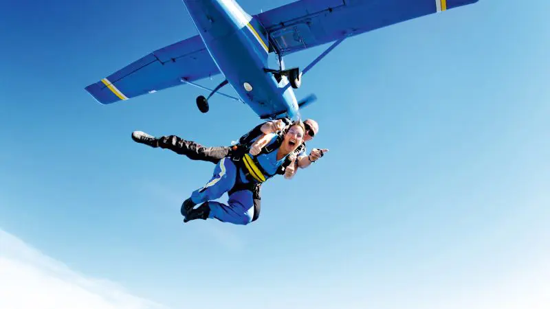 Tandem skydivers soar near Newcastle, freefalling from a blue aeroplane at 15,000ft; one person spreads arms wide with excitement.