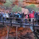 A happy group on a 3-Night Rock adventure smiles and waves from a wooden bridge in a scenic rocky canyon, enjoying the outdoors.